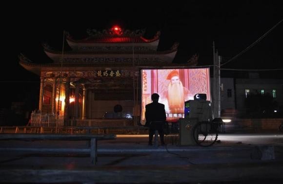 A man sits next to a mobile projector showing a Chinese opera film in front of the local theatre building in the village of Wukan in Lufeng county, Guangdong province December 21, 2011.